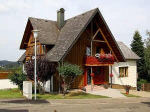 Een houten huis met twee verdiepingen, een flauw hellend dak, een balkon met rode leuningen en een klein voortuintje.