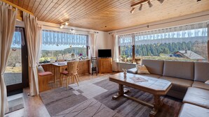 A wooden-paneled room with a sofa, dining table, and chairs, large windows with curtains, and a view of a forested area.