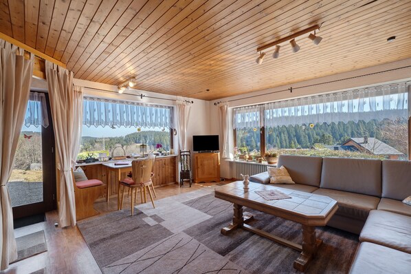 Una habitación con paneles de madera, un sofá, una mesa de comedor y sillas, ventanas amplias con cortinas, y vistas a un área boscosa.