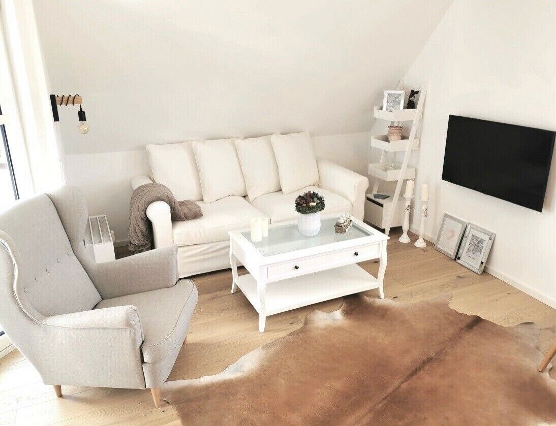 En moderne stue med en hvid sofa, et glasbord, en læder måtte, en hvid lænestol og et ophængt tv.