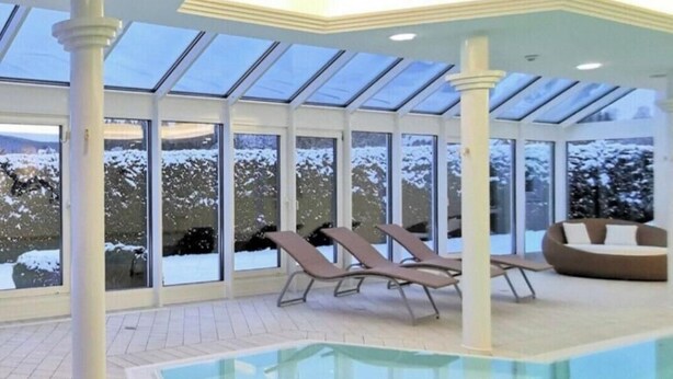 Una piscina interior con techo de vidrio, tumbonas y vistas a árboles cubiertos de nieve.