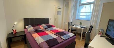 A bedroom with a bed, a desk, a chair, a TV, and a window.