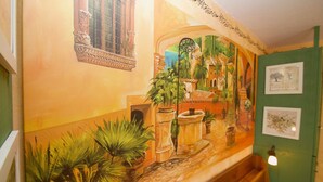 Een kamer met een muurschildering, een raam met decoratief rooster en een groene wand met ingelijste foto's.