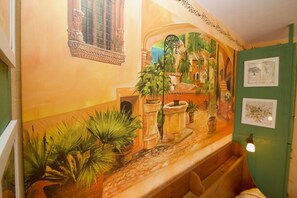 A room with a mural, a window with a decorative grille, and a green wall with framed pictures.