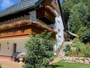 Un chalet traditionnel doté de balcons en bois, d’un escalier en pierre et d’un jardin où poussent diverses plantes et fleurs.
