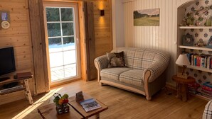 Ein gemütliches Blockhauss-Interieur mit einem Karo-Sofa, Holzwänden, einer Bücherwand und einem großen Fenster.