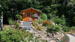 A wooden cabin with stone steps leading up to it, surrounded by lush greenery and flowering plants.