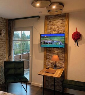Une pièce avec un mur en pierre, un téléviseur fixé au-dessus d’une table en bois, une lampe, un fauteuil vert et un banc.