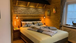Une chambre avec un mur en pierre, des poutres en bois au plafond, un lit agrémenté d’oreillers à motifs, des lampes de chevet et une fenêtre décorée d’une image de cerf.