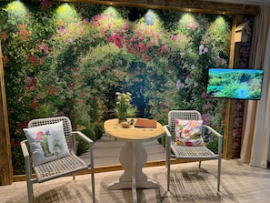 Una habitación con una mesa redonda, dos sillas de mimbre, un televisor en la pared y un mural con motivos florales.