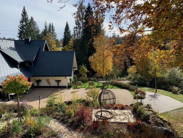 Una casa con tejado inclinado, un jardín con diversas plantas y una escultura metálica esférica.