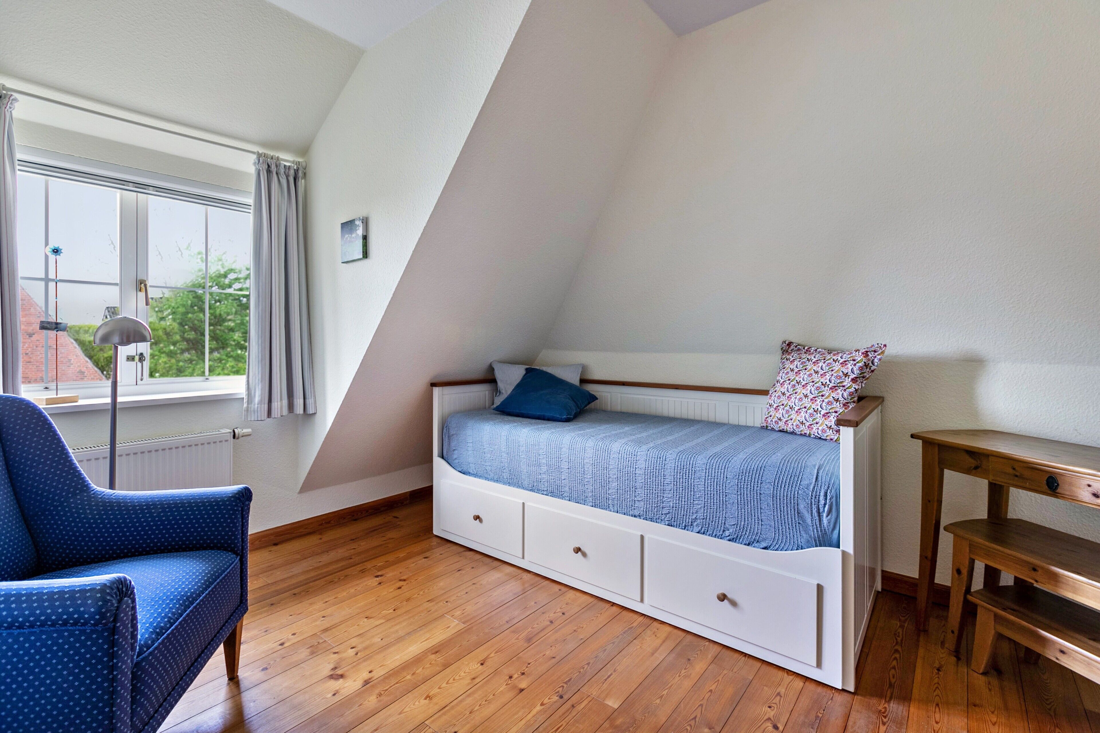 A bedroom with a sloped ceiling, a bed with drawers, a chair, a wooden table, and a window with curtains.
