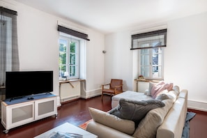 A living room with a television on a cabinet, a sofa with cushions, a chair, and two windows with blinds.