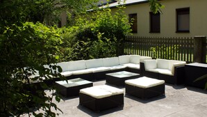 Een terras met een bank en poefs, omgeven door groen en een gebouw op de achtergrond.