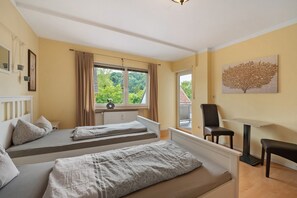 A room with two beds, a table, a chair, a window with curtains, and a painting on the wall.