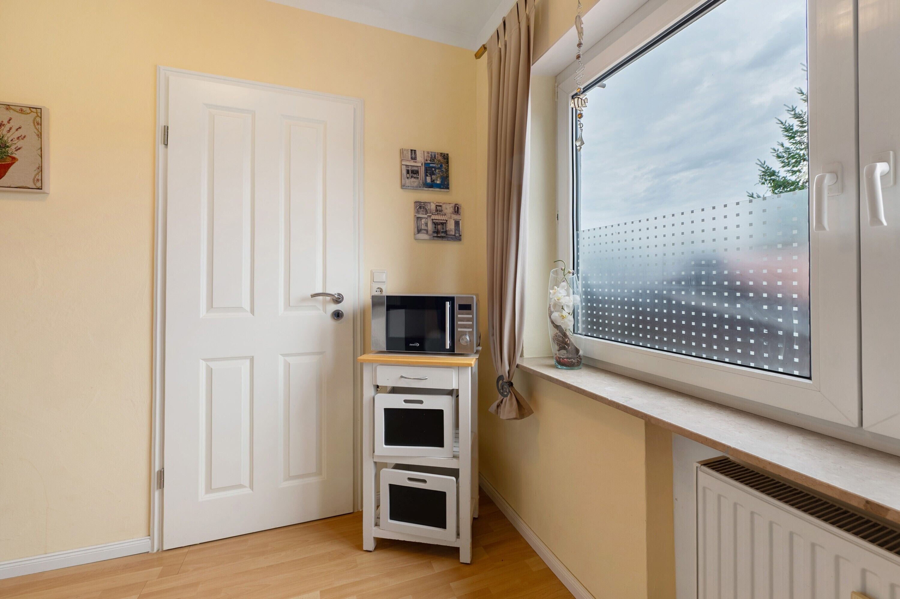 A room with a white door, a window with a view, a microwave on a small white cabinet, and a thermostat on the wall.