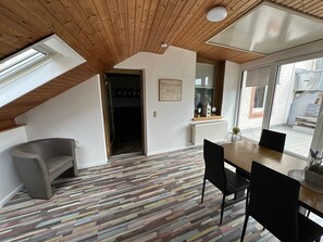Ein Zimmer mit schrägverdeckter Decke, einem Esstisch mit Stühlen, einem gemusterten Boden und einem Fenster mit Aussicht.