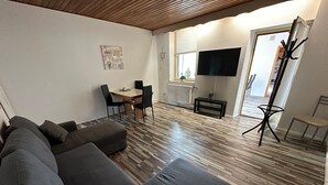A modern living room with a sectional sofa, a dining area with a table and chairs, a flat-screen TV mounted on the wall, and a wooden floor.
