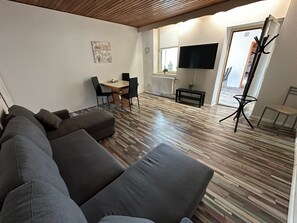 Un salon moderne avec un canapé d’angle, un coin repas comprenant une table et des chaises, un téléviseur à écran plat fixé au mur et un plancher en bois.