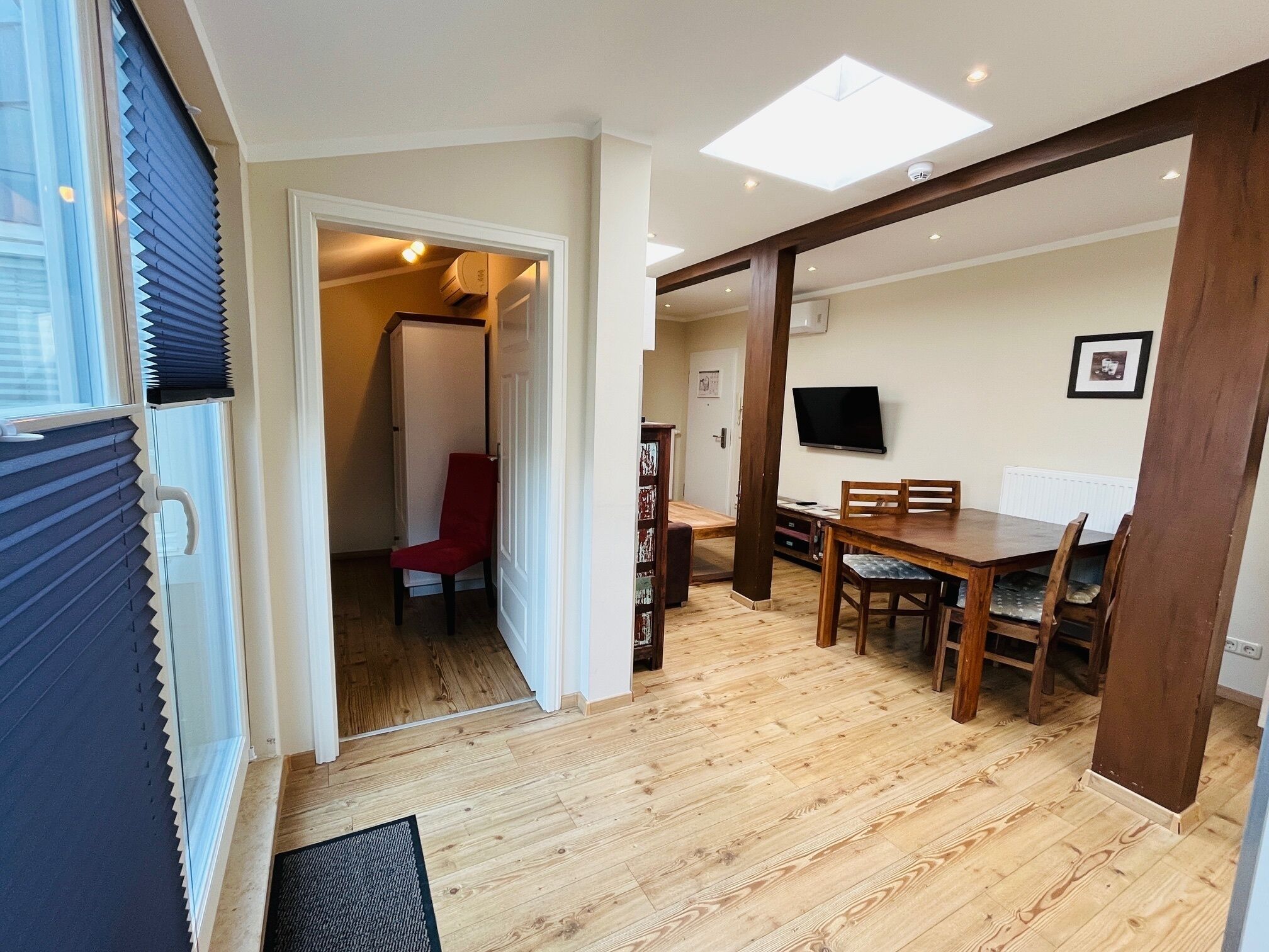 Een eetgedeelte met een houten tafel en stoelen, een rode stoel, een televisie aan de muur en een deur naar een andere kamer.
