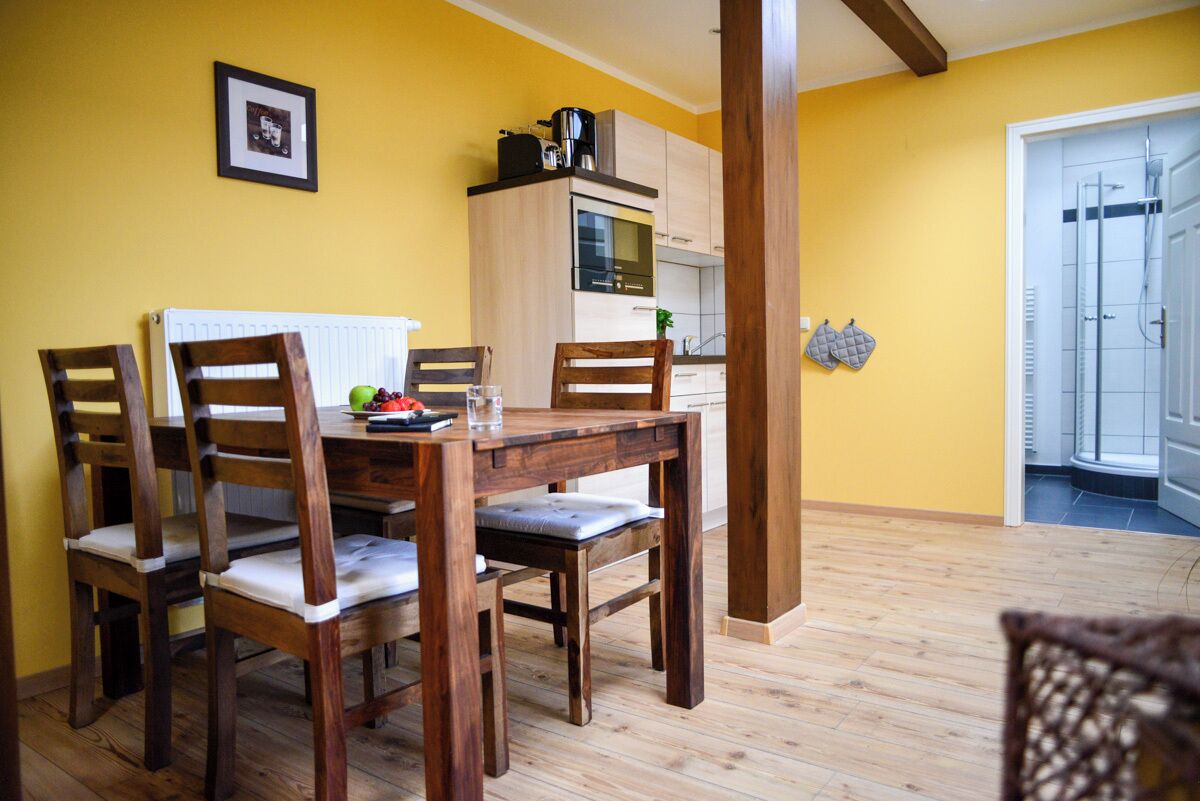 A dining area with a wooden table and chairs, a kitchen in the background, and a yellow wall.
