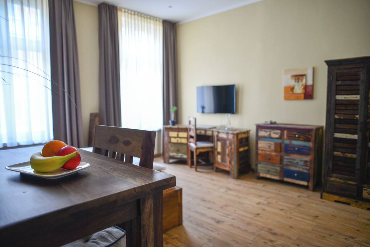 A dining area with a wooden table, chairs, and a fruit plate. There is a television mounted on the wall and a cabinet with drawers.