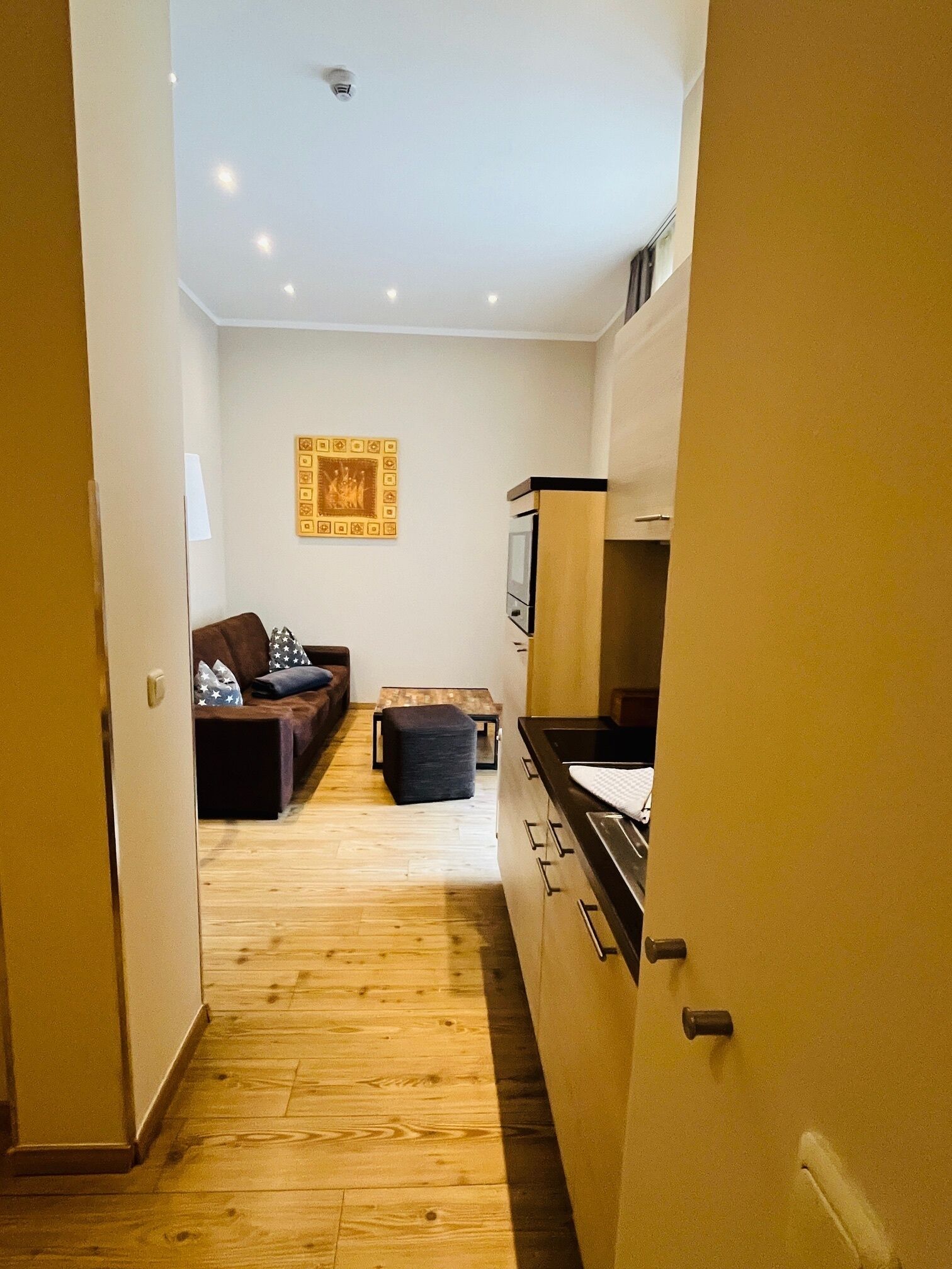 A narrow hallway leading to a living area with a sofa and ottoman, a small table, and a kitchen area with cabinets and a sink.