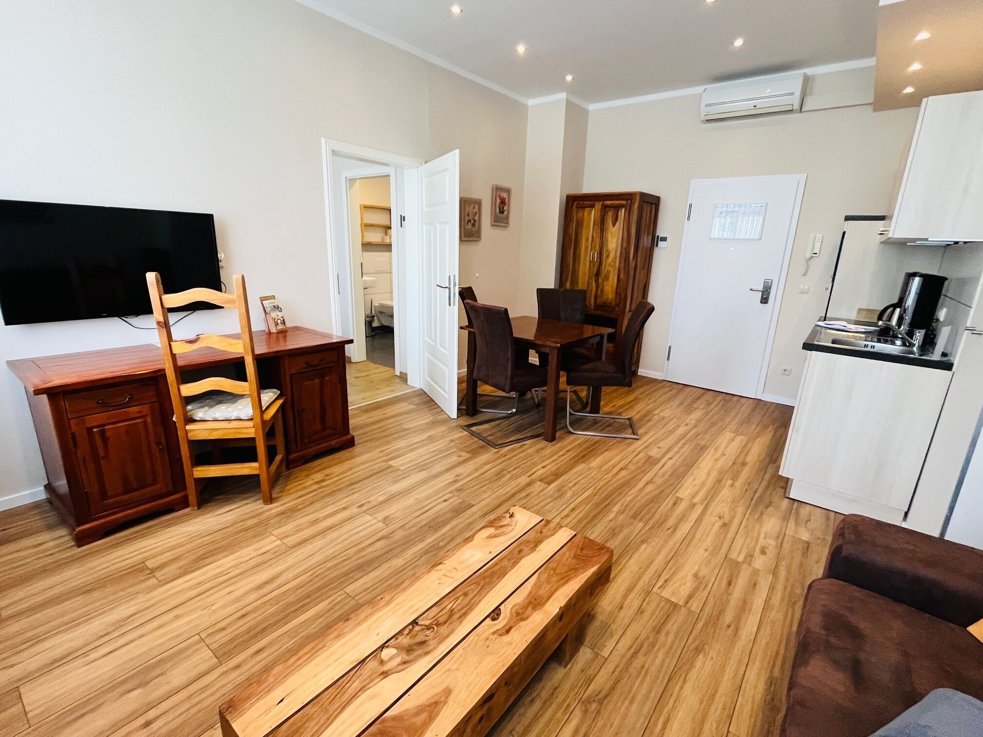 A compact living space with a wooden floor, a dining table with chairs, a bench, a TV mounted on the wall, and a kitchenette area.