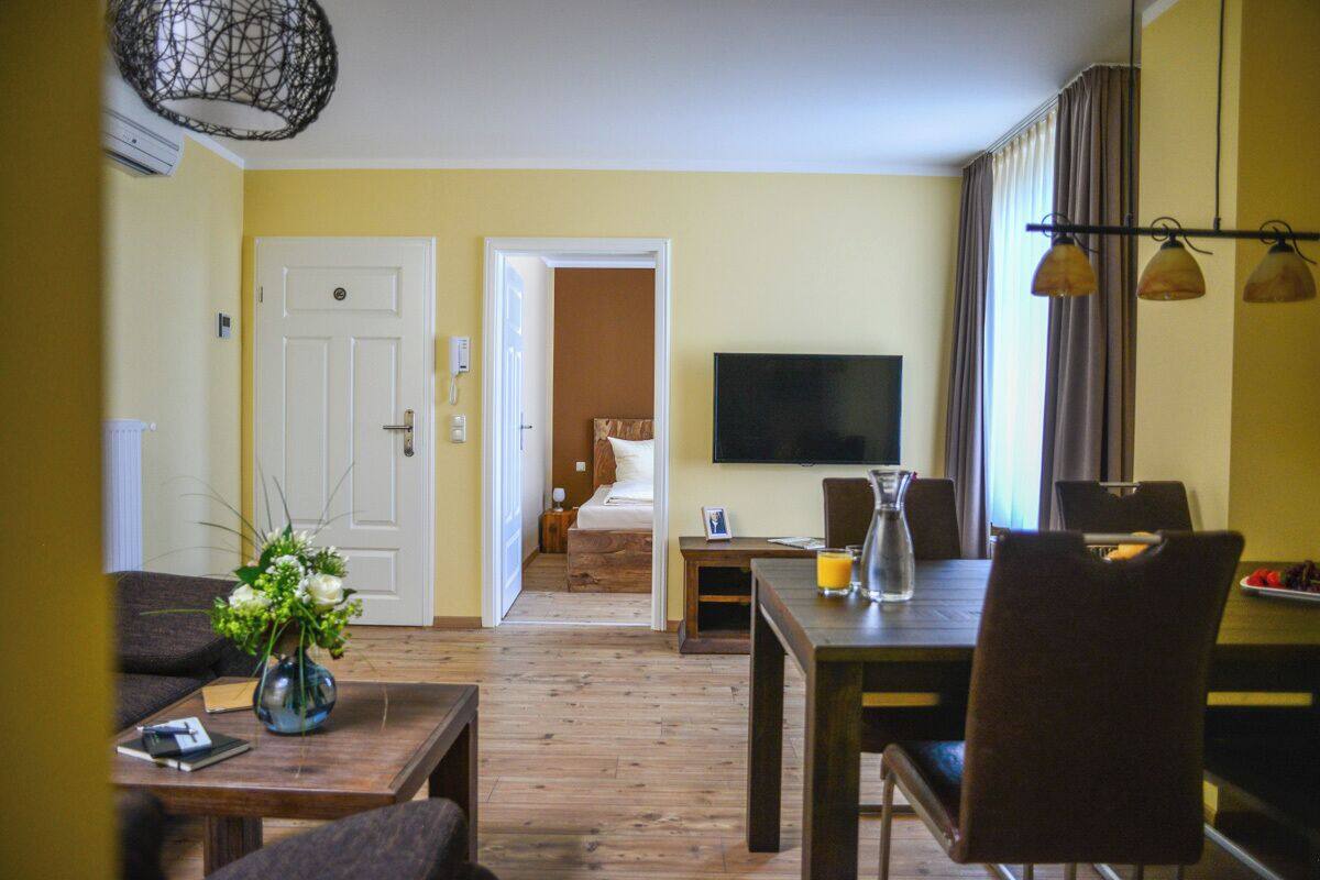 A hotel room with a wooden floor, a dining table with chairs, a TV mounted on the wall, and a bed in the adjacent room.