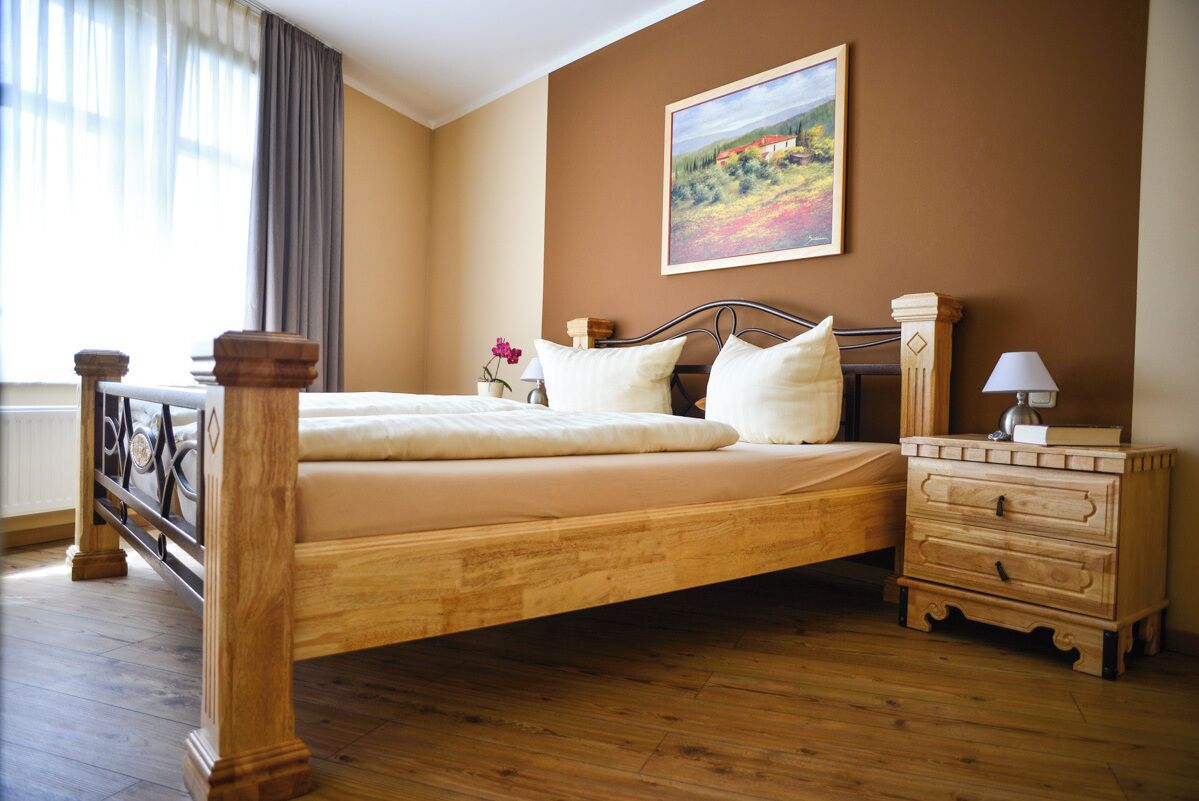 A bedroom with a wooden bed, a nightstand, a dresser, a painting on the wall, and a window with curtains.