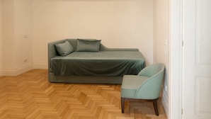 Een kleine, verzorgde kamer met een bank en een stoel, een houten visgraatvloer en witte wanden.