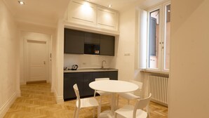 Een kleine keuken met een witte eettafel en stoelen, een inbouwoven en een raam met zonwering.