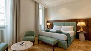 Een hotelkamer met een bed, twee nachtkastjes met lampen, een groene bank, een ronde salontafel en uitzicht op de stad door grote ramen.