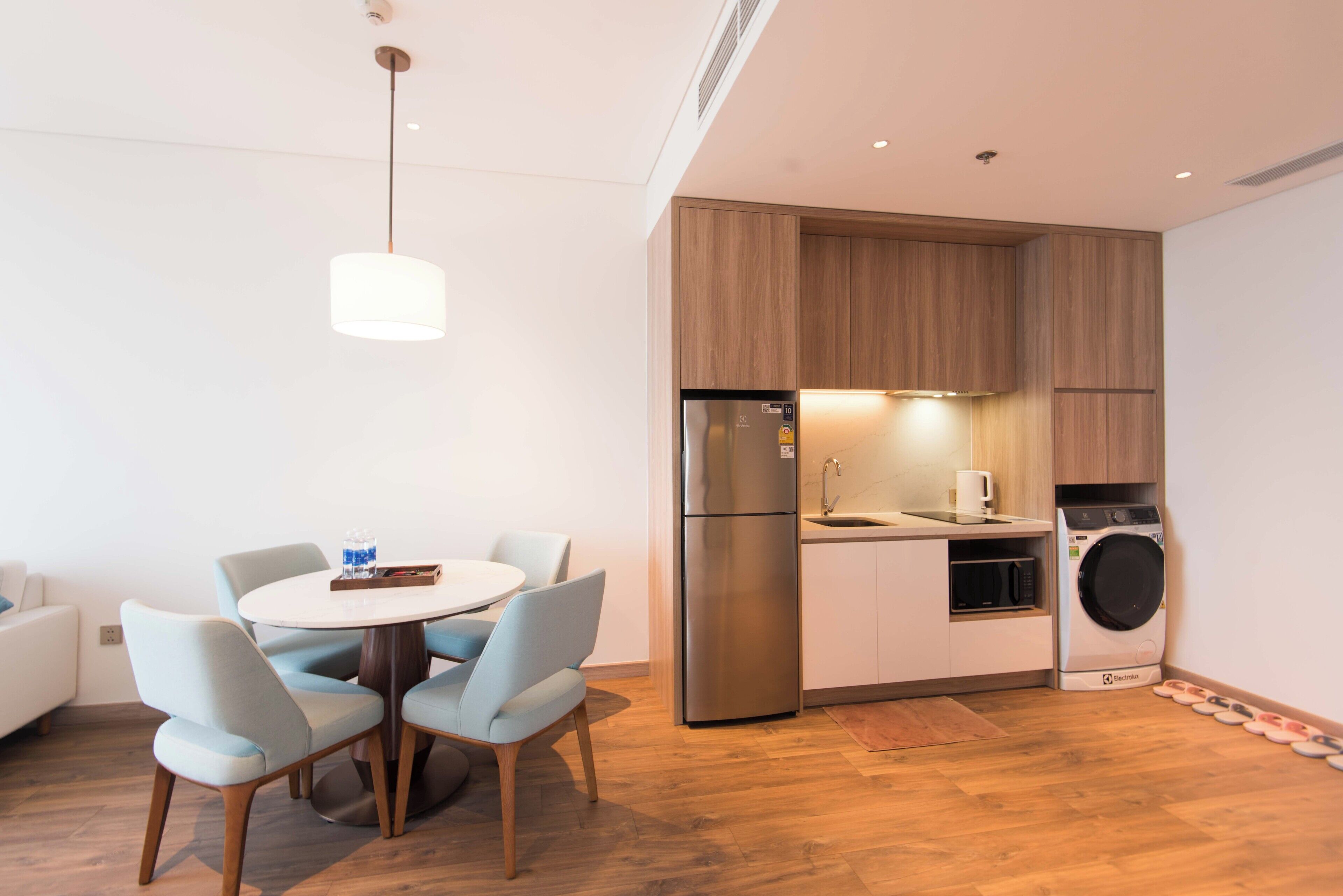 A compact kitchen with a built-in washing machine, a dining area with a round table and chairs, and a hanging light fixture.