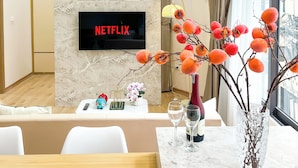 Una sala moderna con un televisor de pantalla plana mostrando 'NETFLIX', una pared de mármol, un mueble de madera y un comedor con una mesa preparada para dos.