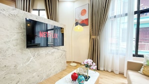 Un moderno salón con una televisión de pantalla plana mostrando 'NETFLIX', una pared de mármol, una mesa de centro con objetos decorativos y un amplio ventanal con cortinas.