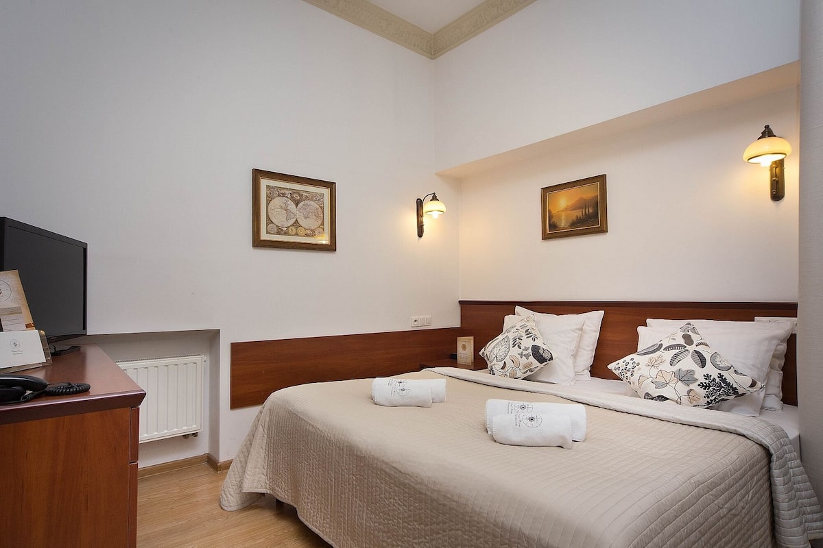 Quarto de hotel com cama, escrivaninha, televisão e duas luminárias fixadas na parede.