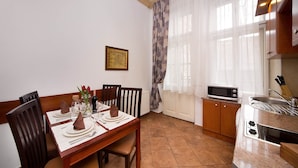 Un comedor con una mesa de madera preparada para cuatro, una cocina equipada con microondas y tostadora, y un ventanal con cortinas traslúcidas.