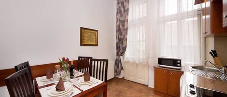 A dining area with a wooden table set for four, a kitchen with a microwave and toaster, and a window with sheer curtains.