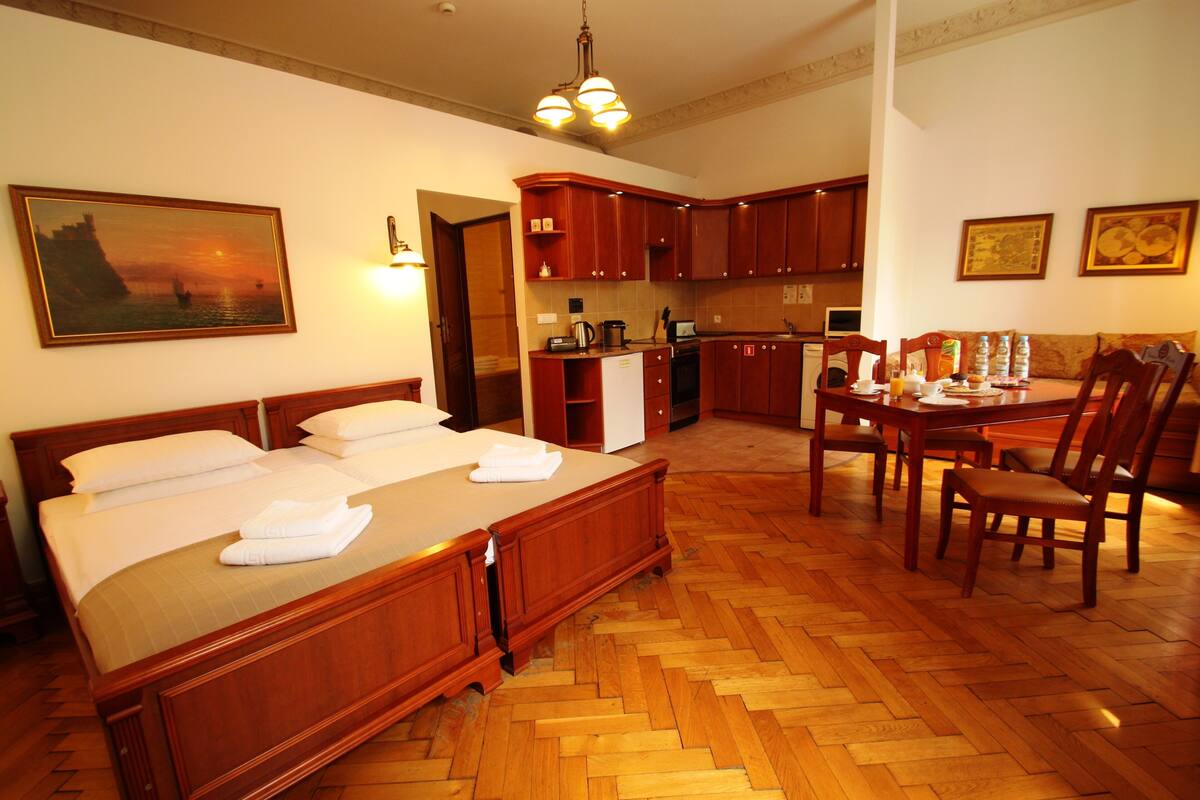 Quarto de hotel com cama, piso de madeira, mesa de jantar com cadeiras, cozinha e obras de arte nas paredes.
