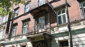 Un edificio de varios pisos con fachada de ladrillo, un balcón con barandilla de metal ornamentada y una puerta con un cartel arriba.