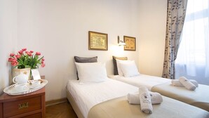 Habitación de hotel con dos camas, una mesita de noche de madera con una bandeja de alimentos para el desayuno, un jarrón de flores rosadas y dos cuadros en la pared.