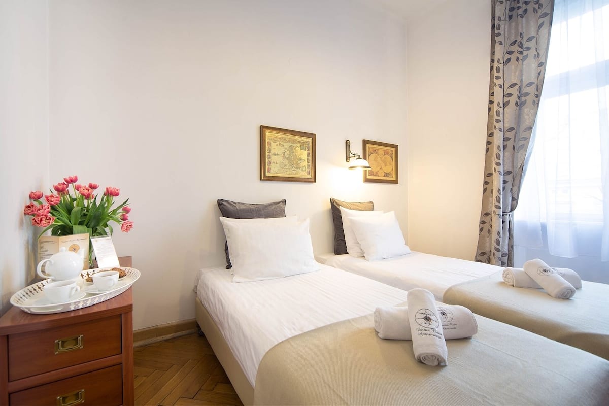 Quarto de hotel com duas camas, uma mesa de cabeceira de madeira com uma bandeja de itens de café da manhã, um vaso de flores cor-de-rosa e duas fotos emolduradas na parede.
