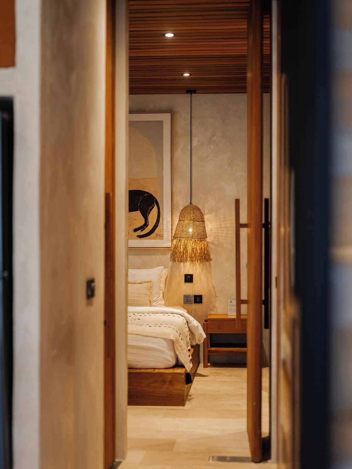 A narrow hallway leading to a bedroom with a bed, a hanging lamp, and a framed artwork on the wall.