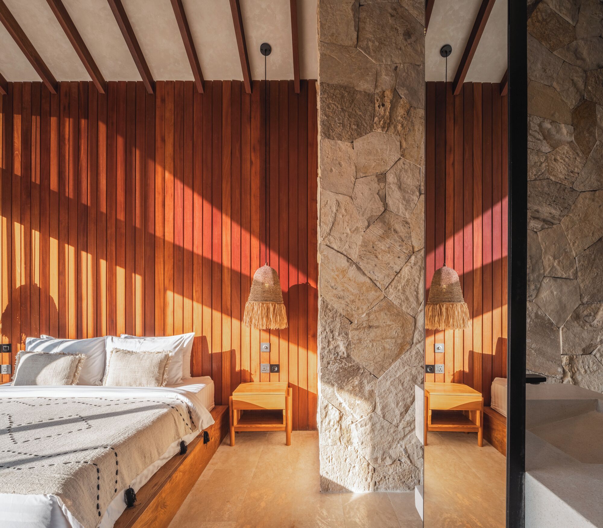 A bedroom with a wooden slatted ceiling, a stone accent wall, a bed with white bedding, and a small wooden table with a hanging lamp.
