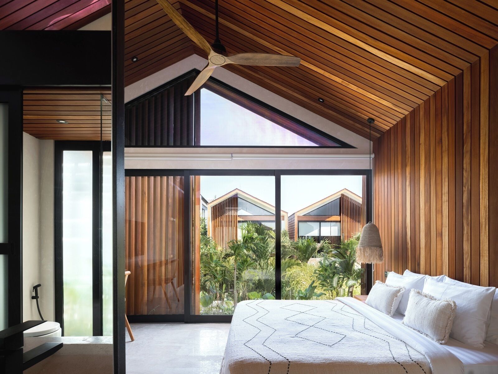 A modern bedroom with a large bed, a ceiling fan, and a view of a residential area through large windows.