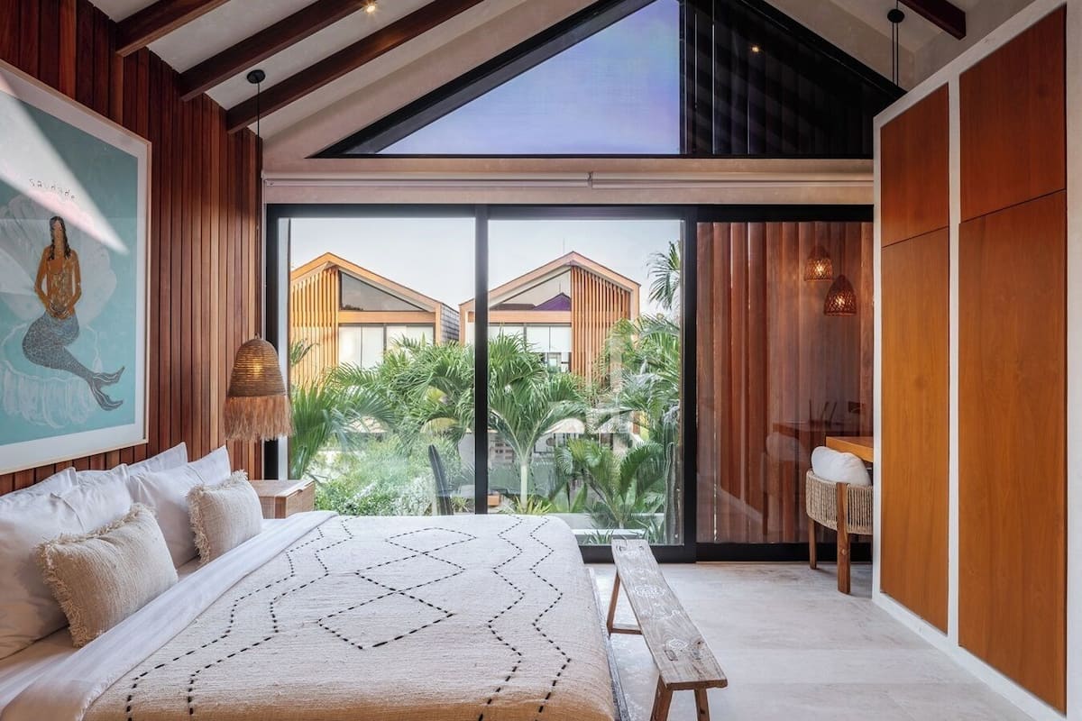 A modern bedroom with a large bed, wooden ceiling beams, a large window, and a view of a building outside.