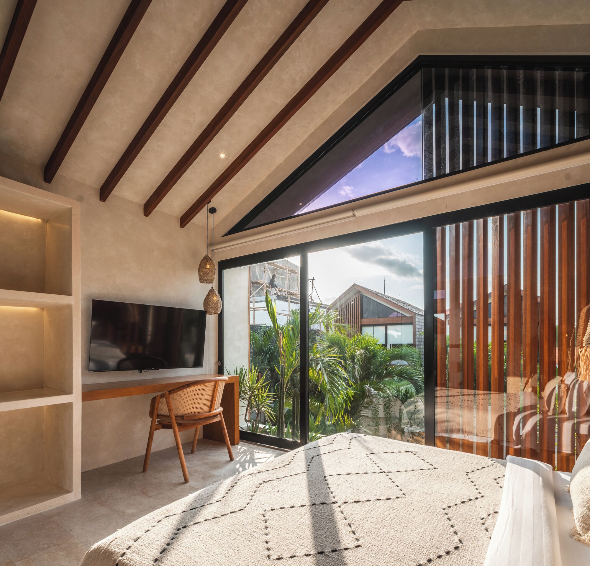 Una habitación moderna con un gran ventanal corredero, techo de madera, televisor montado en la pared y una zona de estar con una silla tejida y una alfombra estampada.