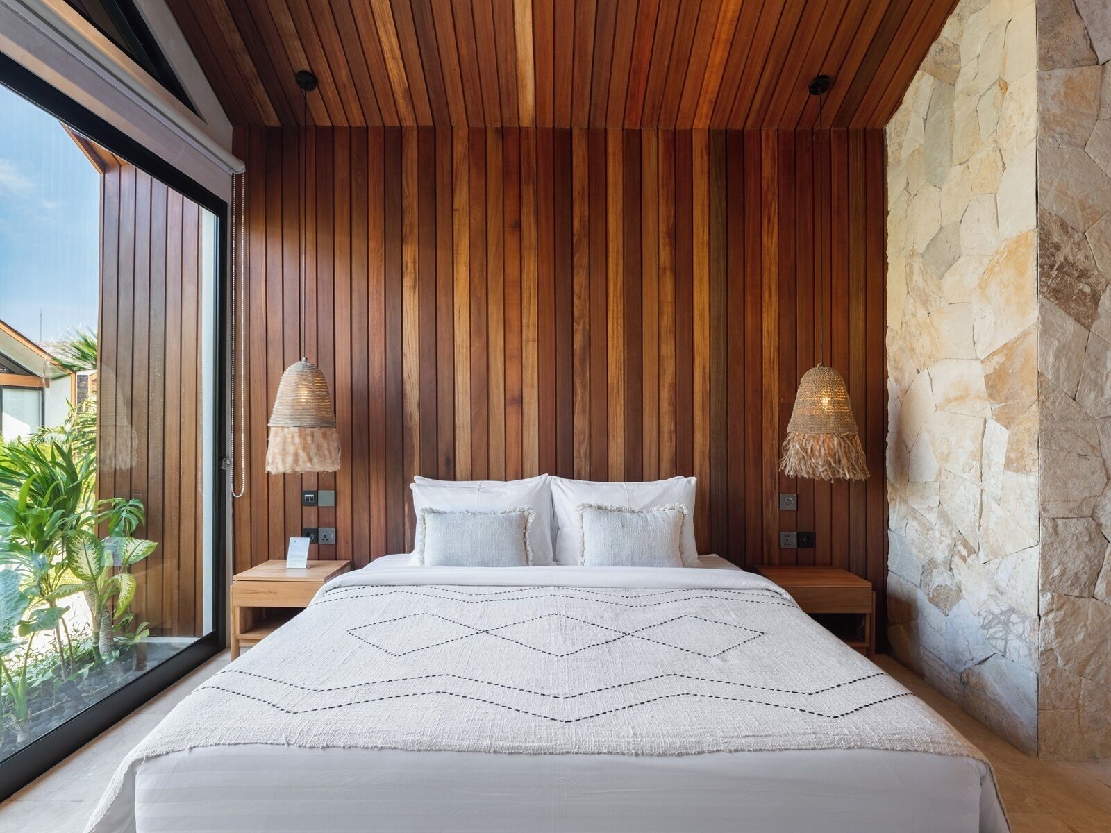 A bedroom with a wooden slatted ceiling, a stone accent wall, a large bed with a white bedspread, and two bedside lamps.