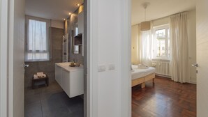 Une salle de bain moderne avec une douche, un lavabo et un miroir. On y trouve un petit banc et un porte-serviettes. La salle de bain est carrelée et possède une fenêtre avec des rideaux.